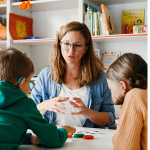 Eine Frau kommuniziert an einem Tisch mit zwei Kindern, die Hörsysteme tragen, und spielt mit den Kindern ein Brettspiel
