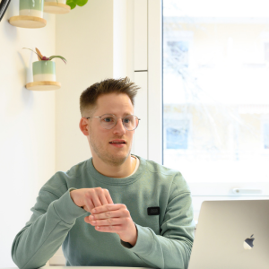 Ein junger Mann mit Brille sitzt an einem Schreibtisch mit Laptop und gebärdet.