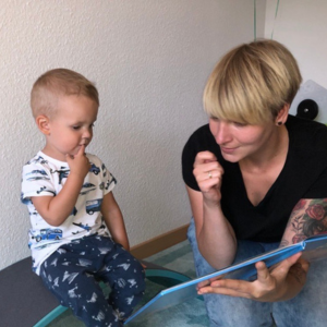 Eine Frau mit kurzen blonden Haaren hält ein aufgeschlagenes Bilderbuch und gebärdet. Ihr gegenüber sitzt ein Kleinkind und führt die Hand zum Kinn, um die Gebärde nachzumachen.