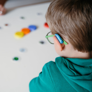 Nahaufnahme eines Kindes von hinten: Es trägt eine Brille mit grünen Bügeln und ein Hörgerät hinter dem Ohr. Vor ihm liegen bunte Spielsteine auf einem hellen Tisch.