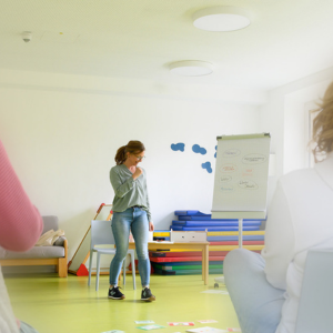 Eine Frau steht in einem hellen Gruppenraum vor einem Flipchart und gebärdet.
