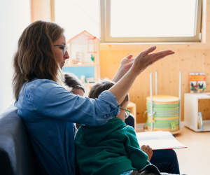 Eine Frau sitzt mit zwei Kindern auf dem Sofa und liest ein Buch vor. Dabei wird eine Gebärde gemacht.