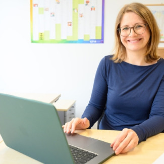 Mitarbeiterin Anna Werle sitzt am Schreibtisch mit ihrem Laptop und lächelt in die Kamera. 