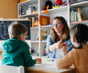 Eine Frau sitzt mit zwei Kindern an einem Tisch und spielt ein Spiel. Dabei wird eine Gebärde gezeigt. 