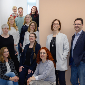 Gruppenfoto von zwölf Mitarbeitenden, die in einem Treppenhaus und vor einer hellen Wand stehen oder sitzen und freundlich in die Kamera lächeln.