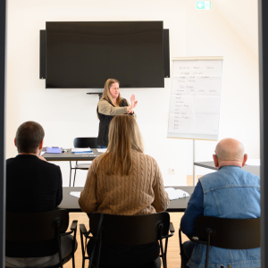 Eine Referentin steht vor einer kleinen Gruppe und hält einen Vortrag. Neben ihr befindet sich ein Flipchart, im Hintergrund hängt ein großer Bildschirm.