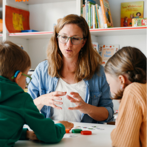 Eine Frau mit Brille und Jeansjacke und gebärdet mit zwei Kindern, die mit dem Rücken zur Kamera sitzen. Im Hintergrund ein Regal mit Büchern und Spielmaterialien.
